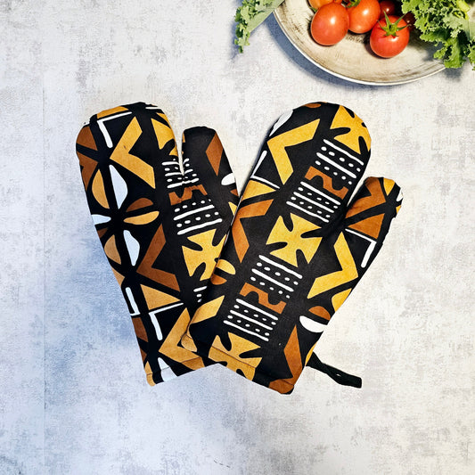 Pair of patterned oven mitts on a textured surface with a bowl of tomatoes in the background.