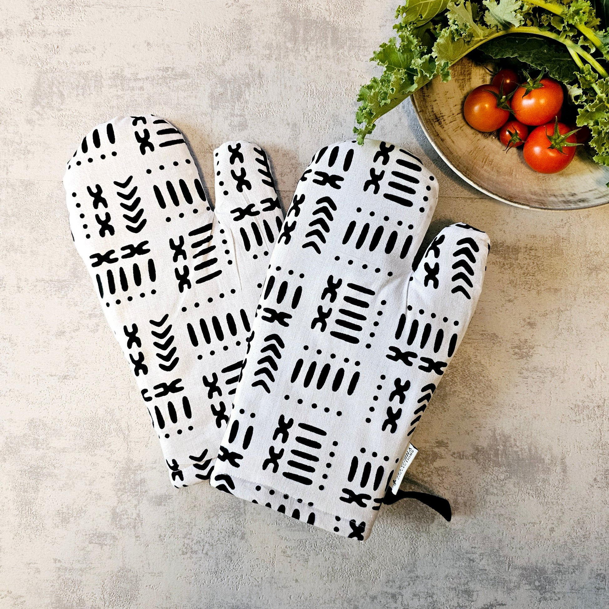 Pair of white oven mitts with black pattern on a neutral surface with a bowl of tomatoes and greens.